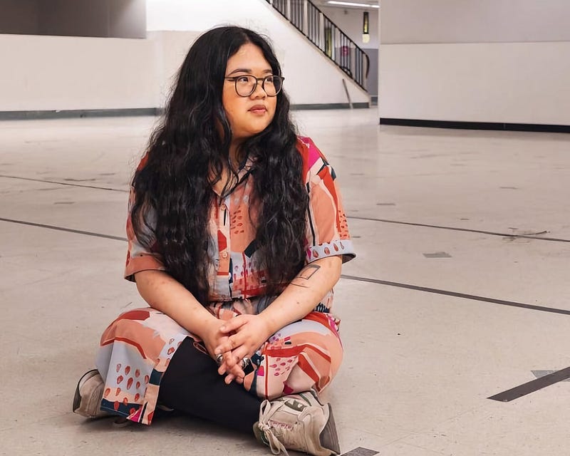 In a bright, minimalist corridor, artist Gianni sits cross-legged on the floor. She’s wearing a vivid, red and orange patterned outfit with black accents and casual sneakers. With long, wavy hair and glasses, she gazes thoughtfully to the side. The corridor, with its modern aesthetic, is marked with directional arrows on the floor.
