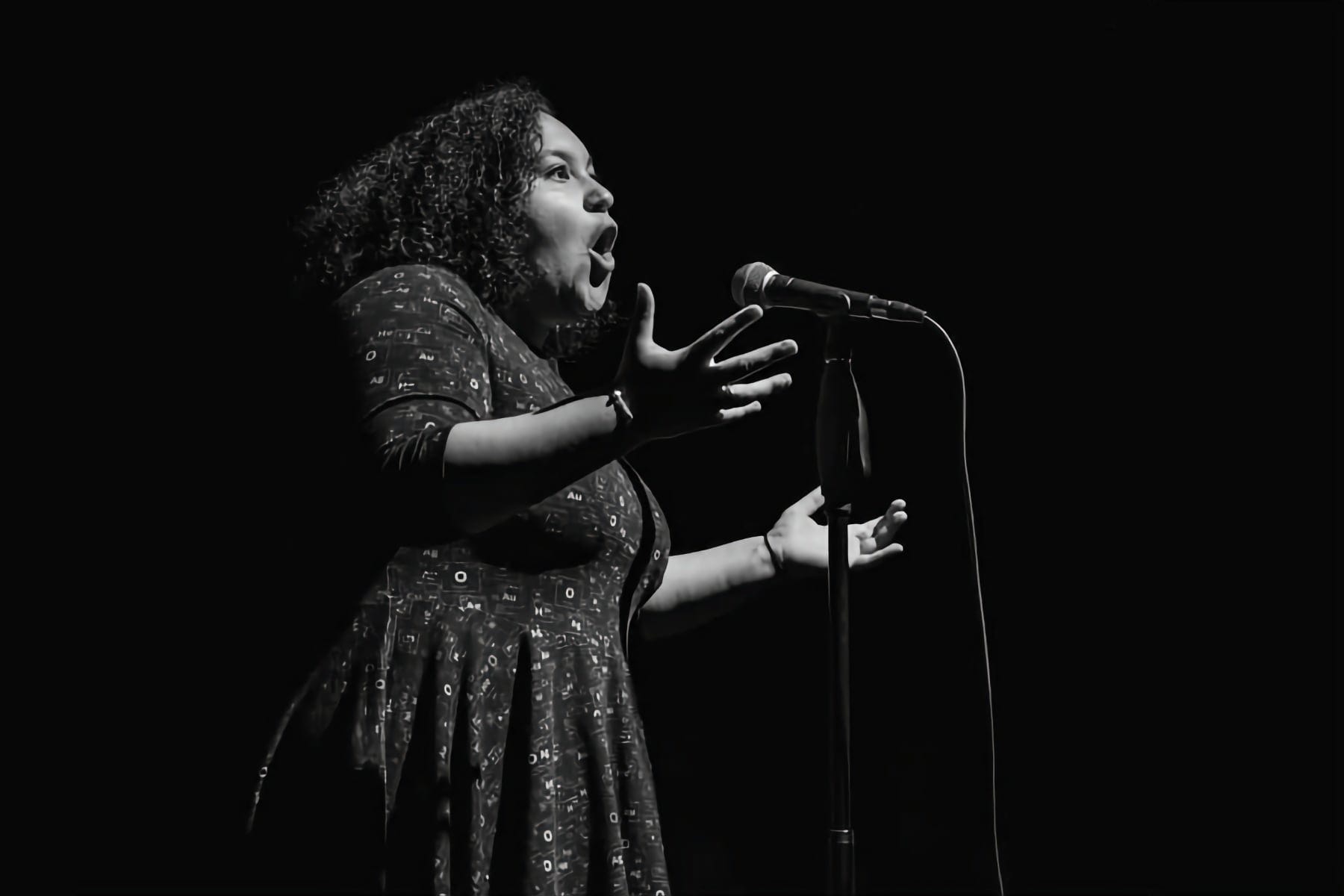 The image is a black and white photograph capturing poet Georgia Bartlett-McNeil in a moment of performance. She is positioned before a microphone, animatedly expressing herself with her mouth open as if caught mid-sentence, hands gesturing to emphasise her spoken word. The lighting casts her in a soft glow against a dark backdrop, highlighting the passion and intensity of her performance.