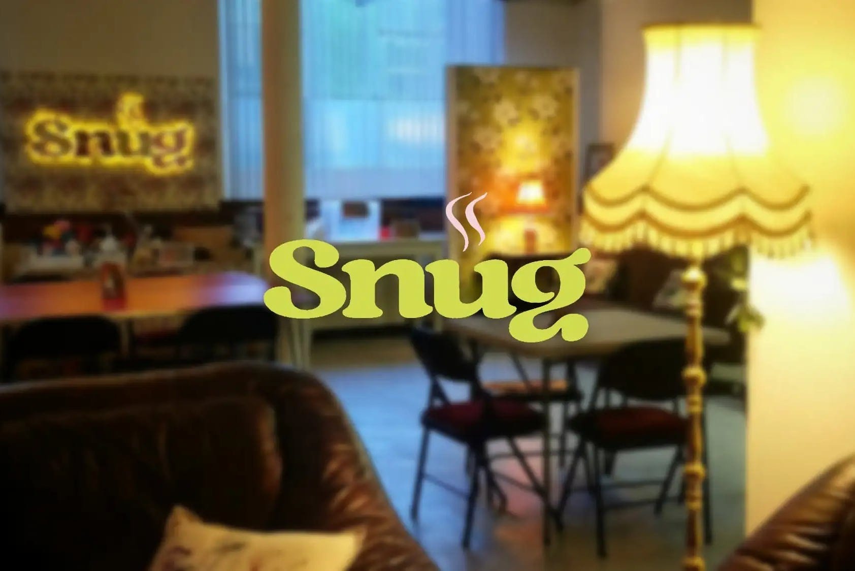 This image shows a cosy, warmly lit room that has an inviting and homely feel. In the foreground, a comfortable-looking brown leather armchair with a cushion invites relaxation. The middle ground reveals a table and chairs, suggesting a dining or social area. A decorative lamp stands to the right, casting a soft glow. The word "Snug," in a large, playful, and bold yellow font with steam lines rising from the 'u', suggests warmth and comfort, overlaying the scene. Behind it, on the back wall, is a blurred neon sign with the same word "Snug," echoing the text in the foreground, enhancing the ambience of the setting.