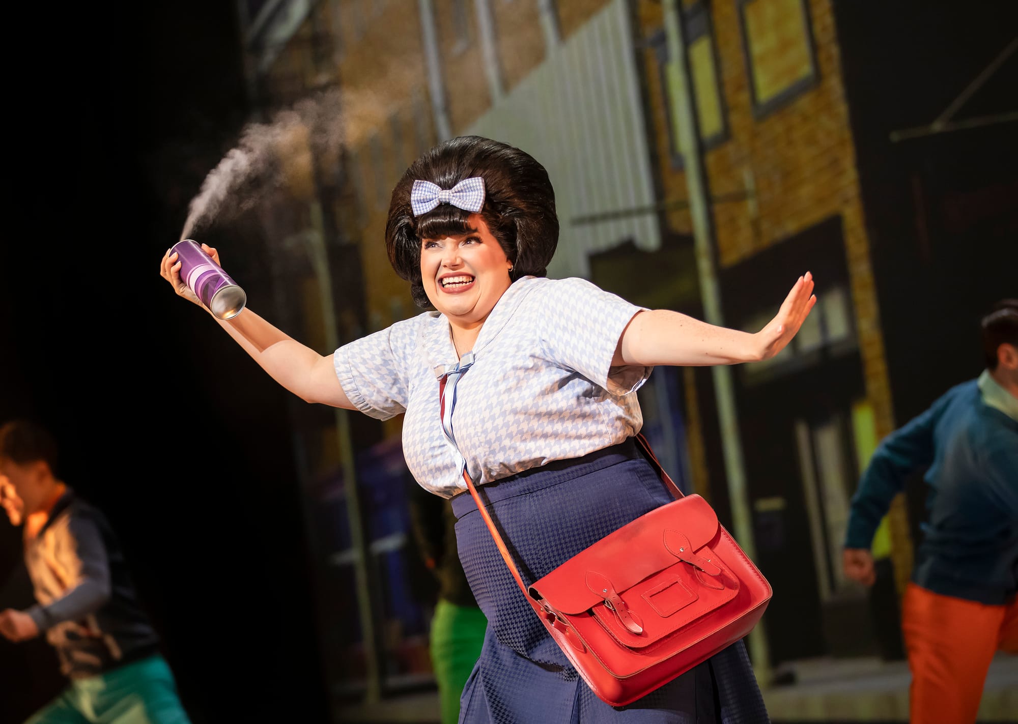 A smiling performer sprays hairspray, dressed in vintage clothing with a red satchel, on a lively stage set.