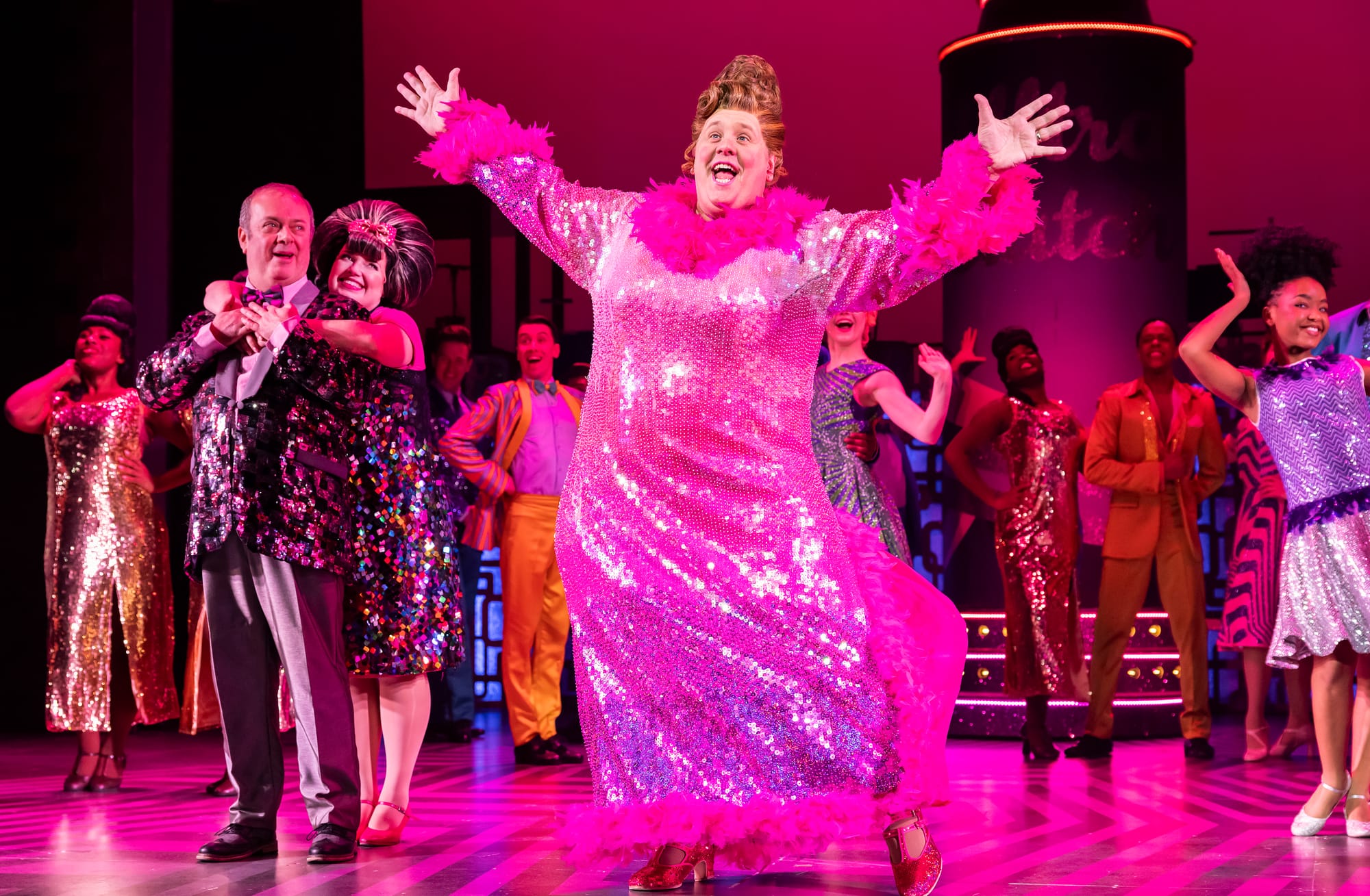 A performer in a sparkling pink gown with cast in retro costumes on a brightly lit musical stage.