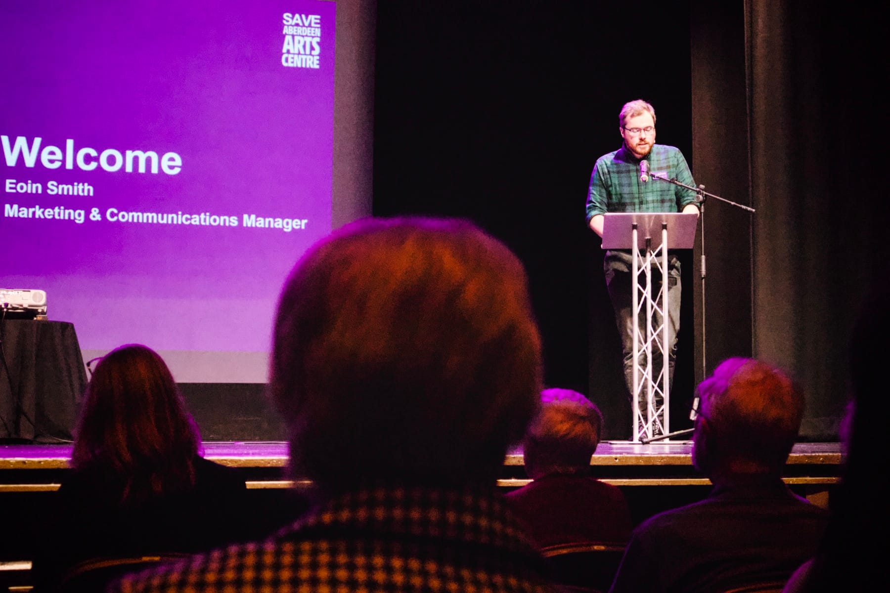 Man in green checkered shirt at a podium, mid-speech, with a purple background screen.