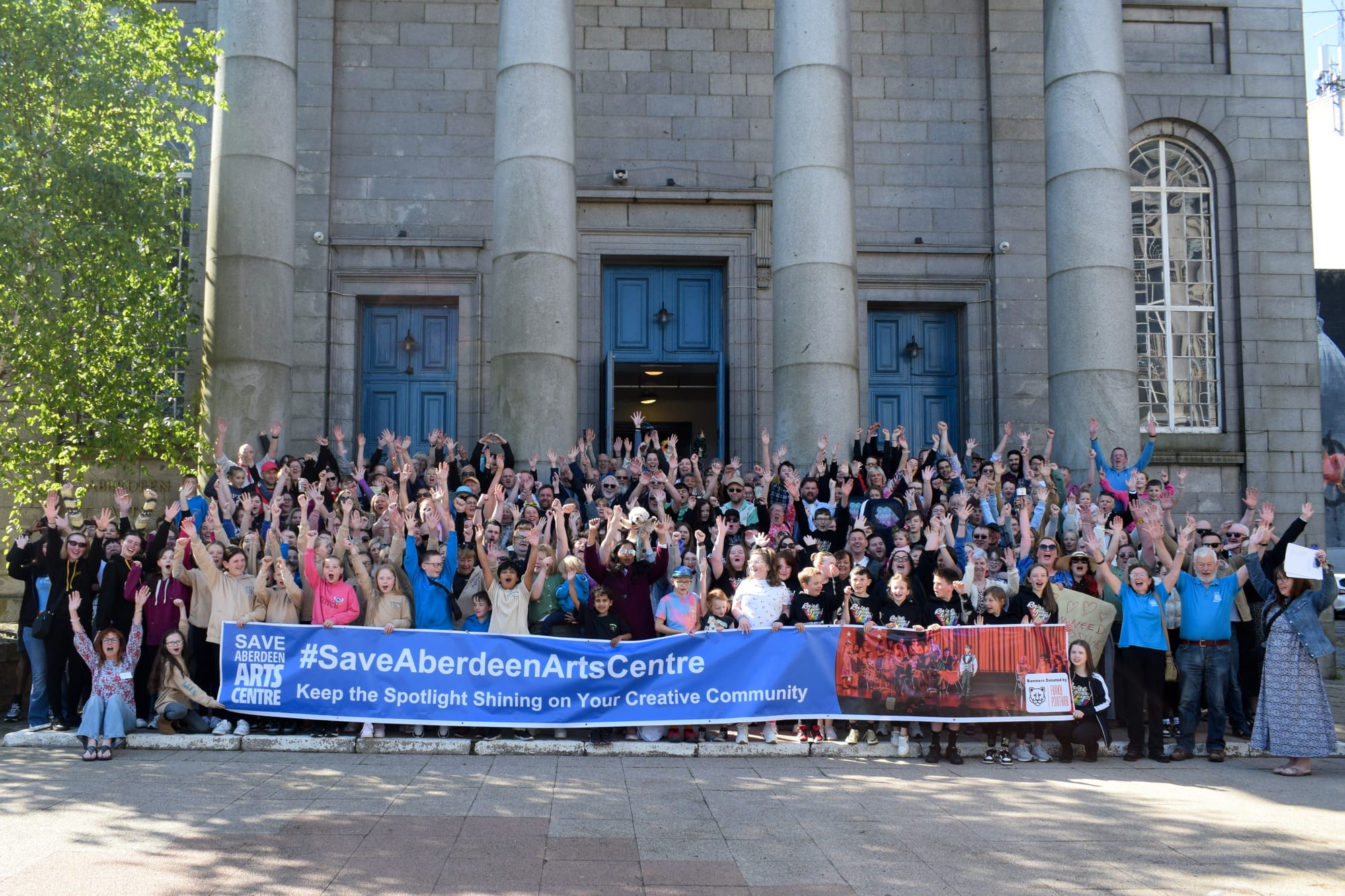 A large, cheerful crowd gathered on the steps of Aberdeen Arts Centre, holding up a “Save Aberdeen Arts Centre” banner and cheering in support of the venue’s future.