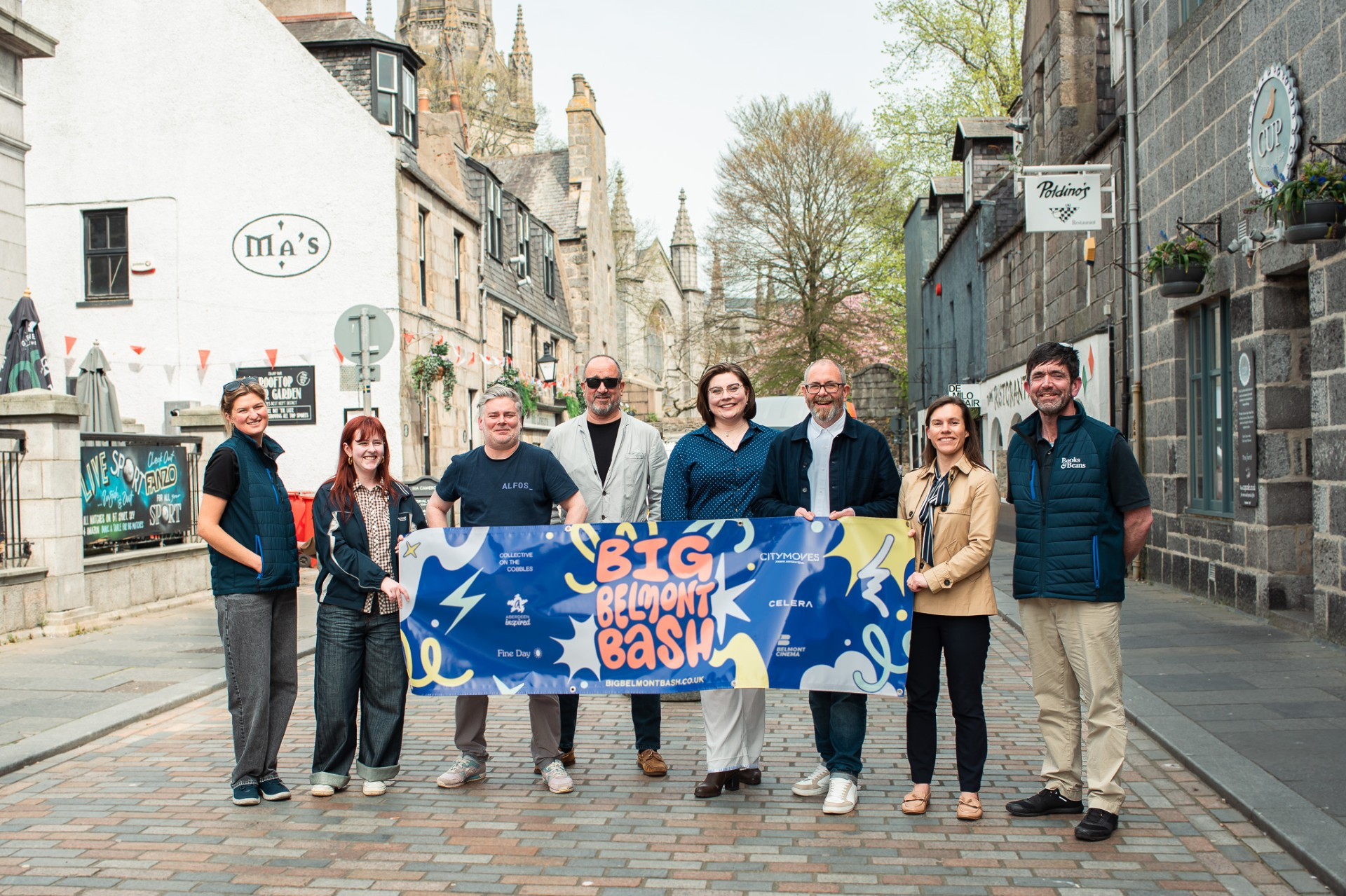 A group of people standing on a cobbled street holding a Big Belmont Bash banner together.