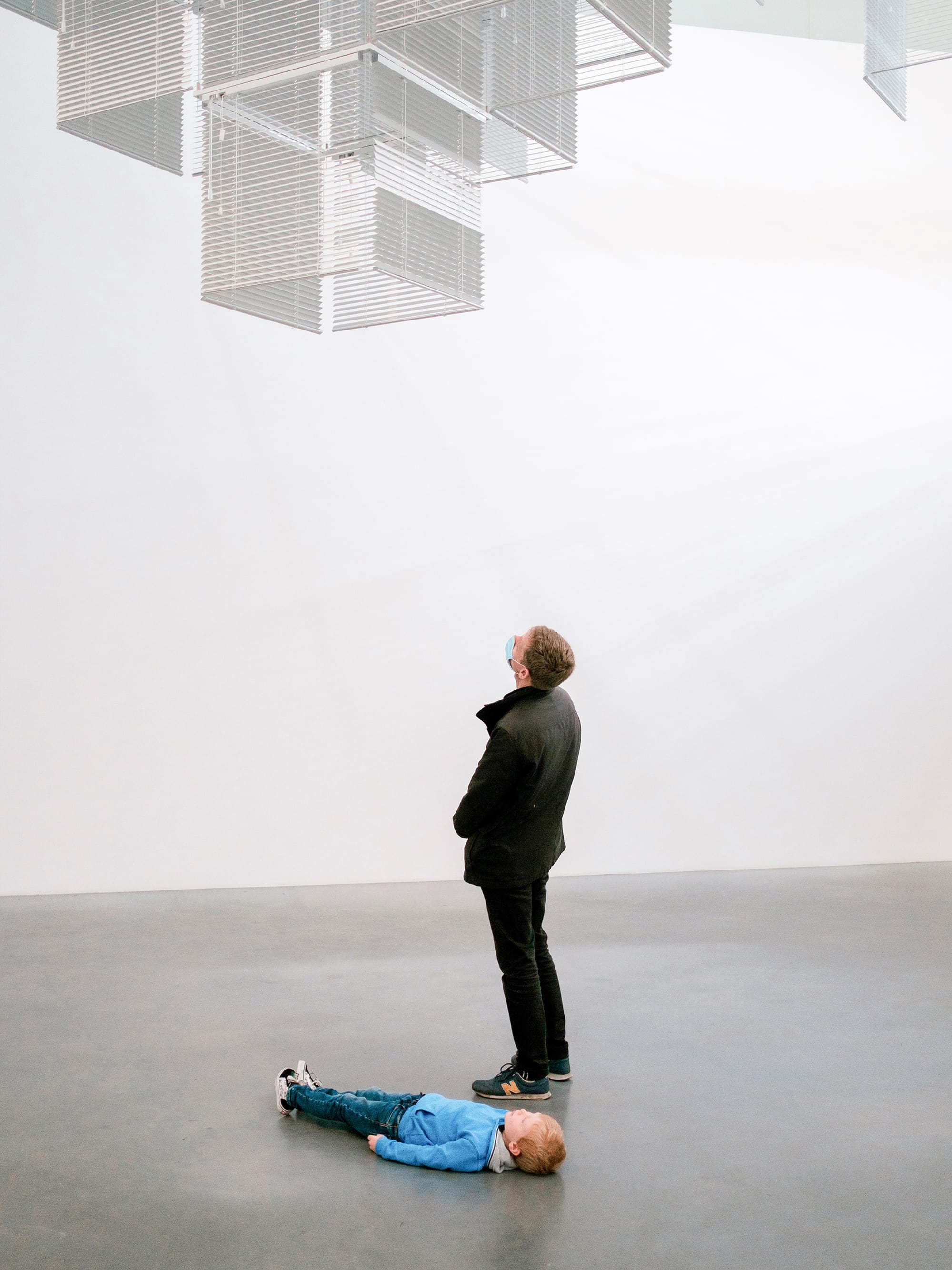 Man stands as child lies beneath cube installation in a white gallery.
