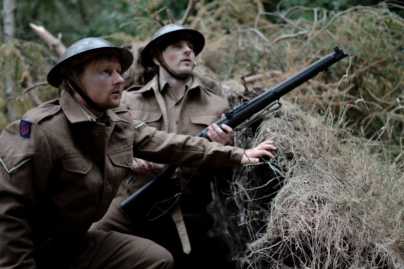 Cameron Prince and Neil Thompson in WWII uniforms crouch in a woodland trench, one holding a rifle.