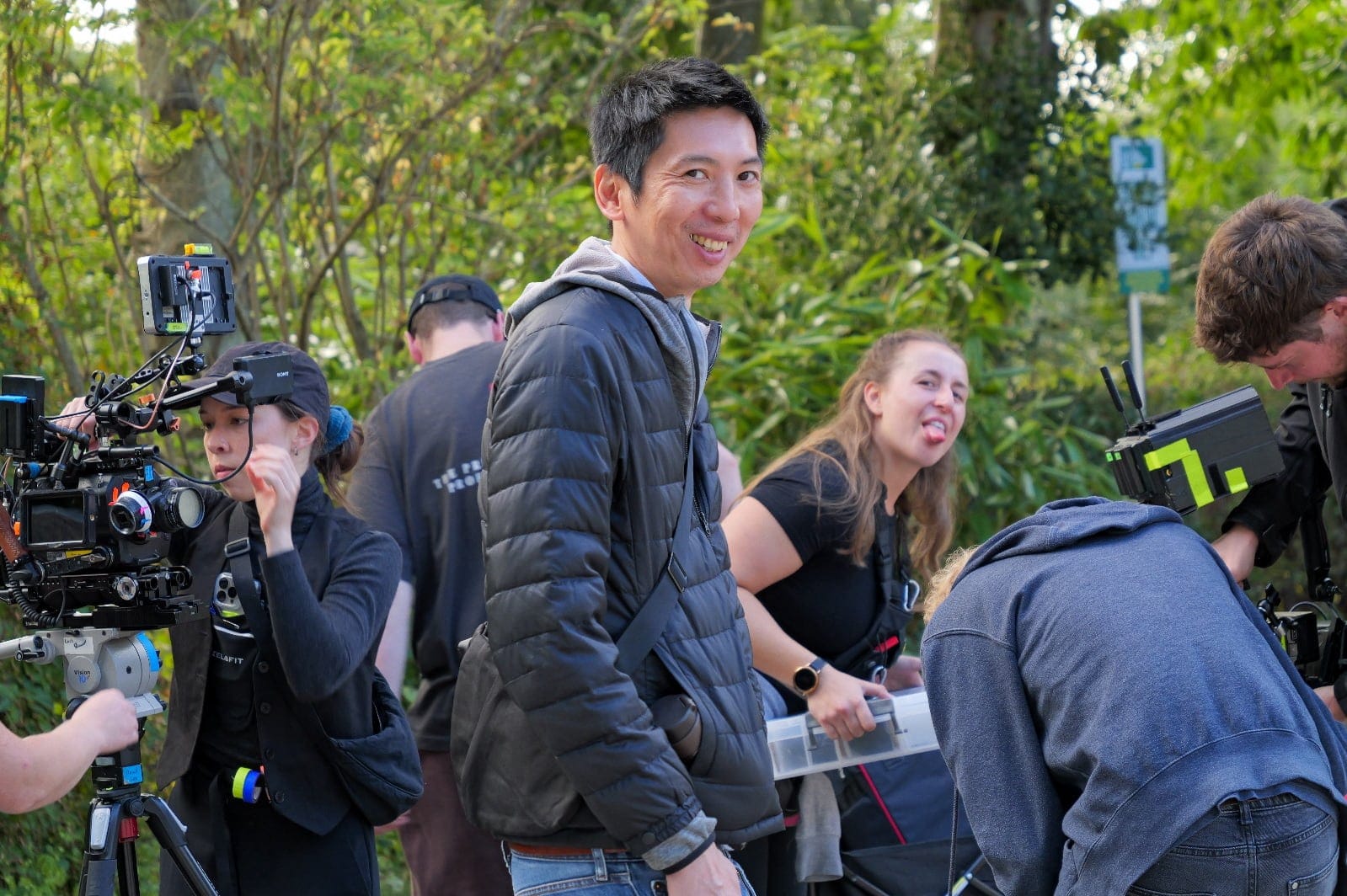 David Lee smiles among crew members adjusting cameras and equipment during an outdoor shoot.