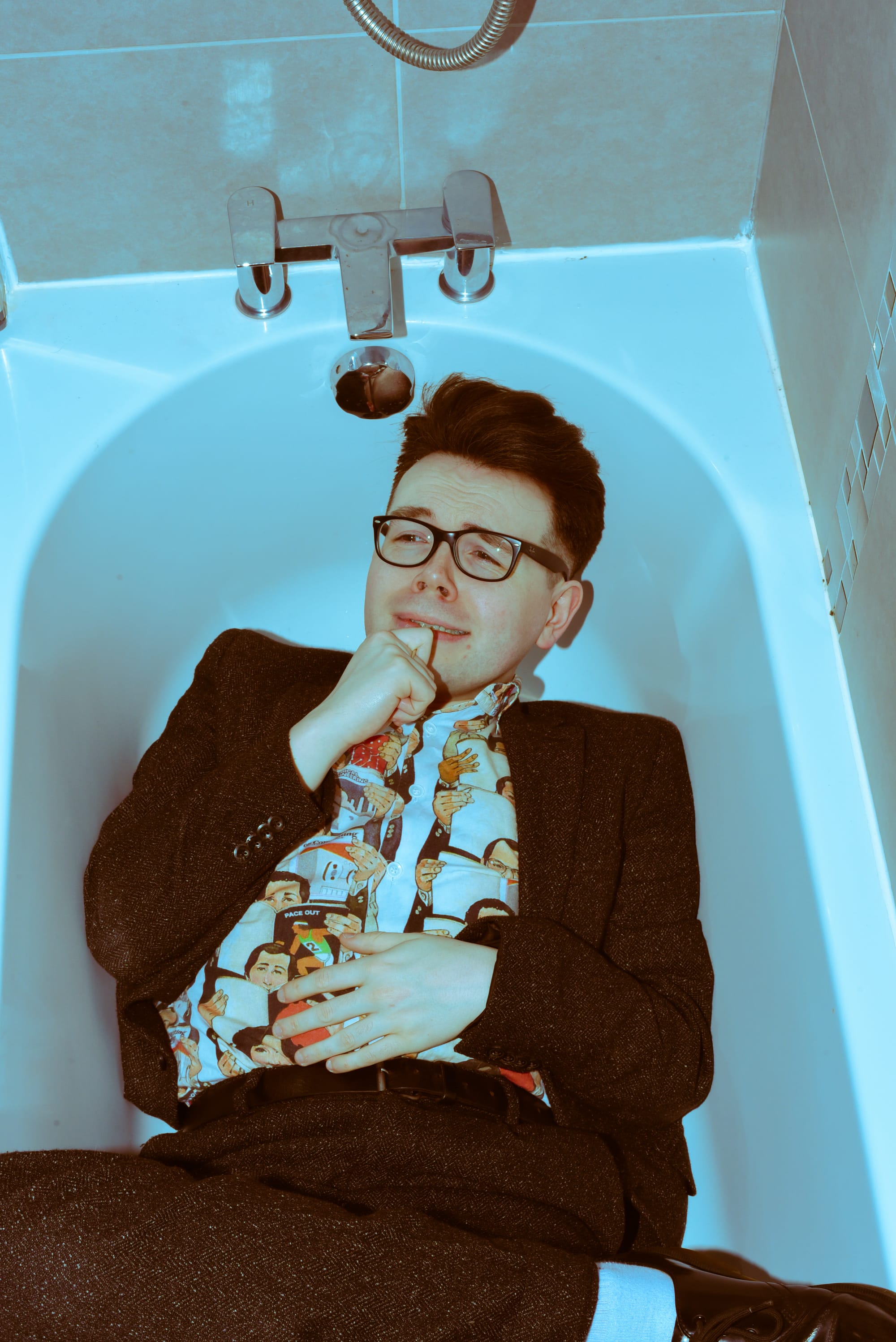 Christopher Macarthur-Boyd in glasses and suit, lying in white bathtub, shot from above