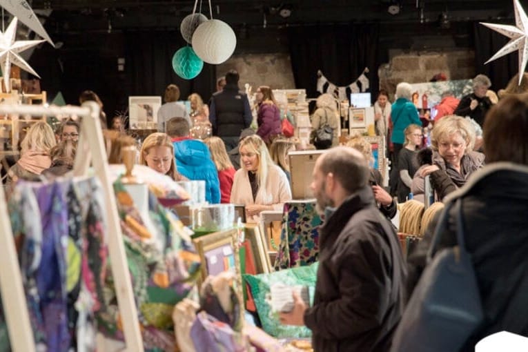 Busy indoor craft fair with visitors browsing stalls displaying artwork and crafts, colourful paper lanterns hanging overhead.