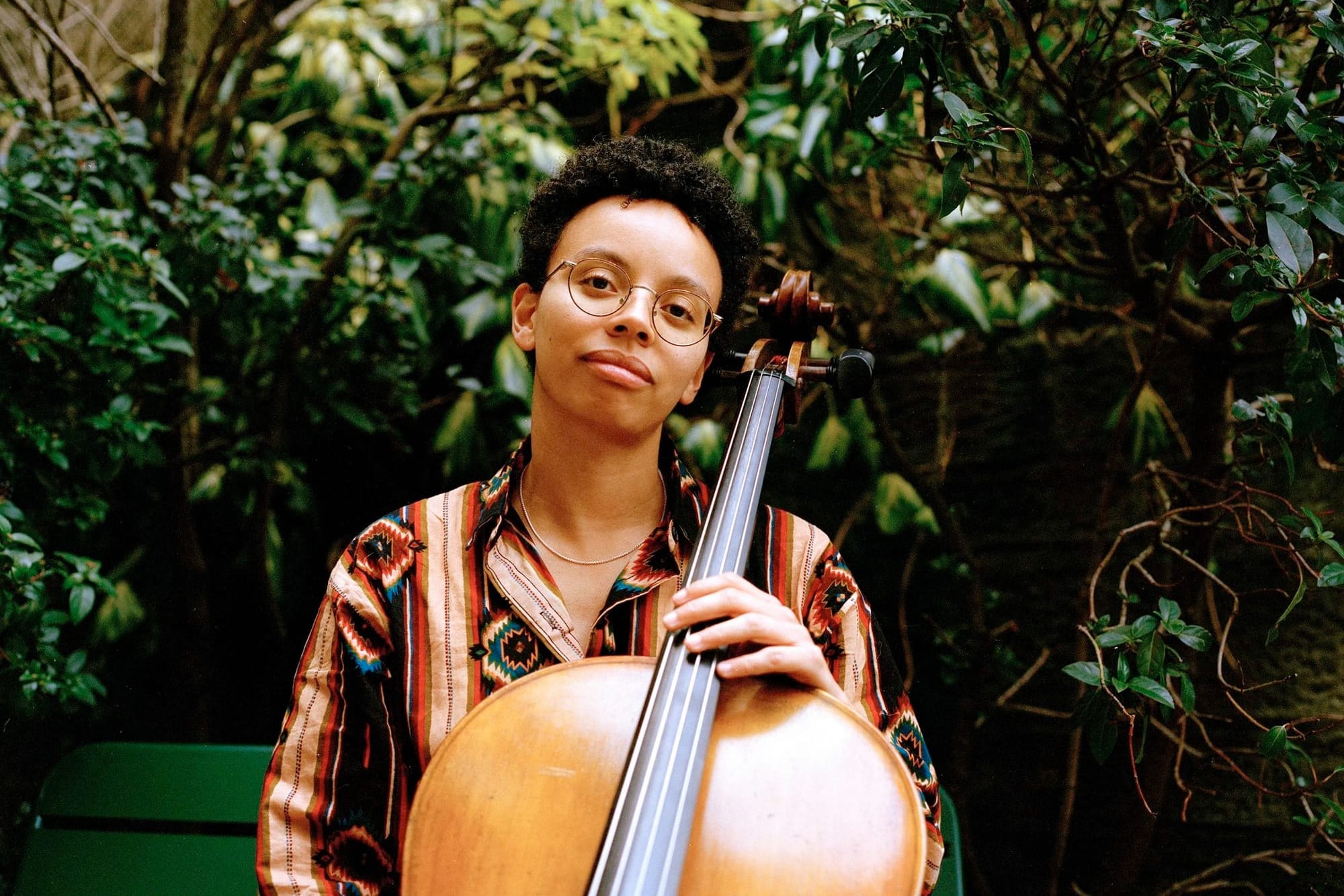 Simone Seales holding cello, wearing glasses and patterned orange shirt, against green foliage background.