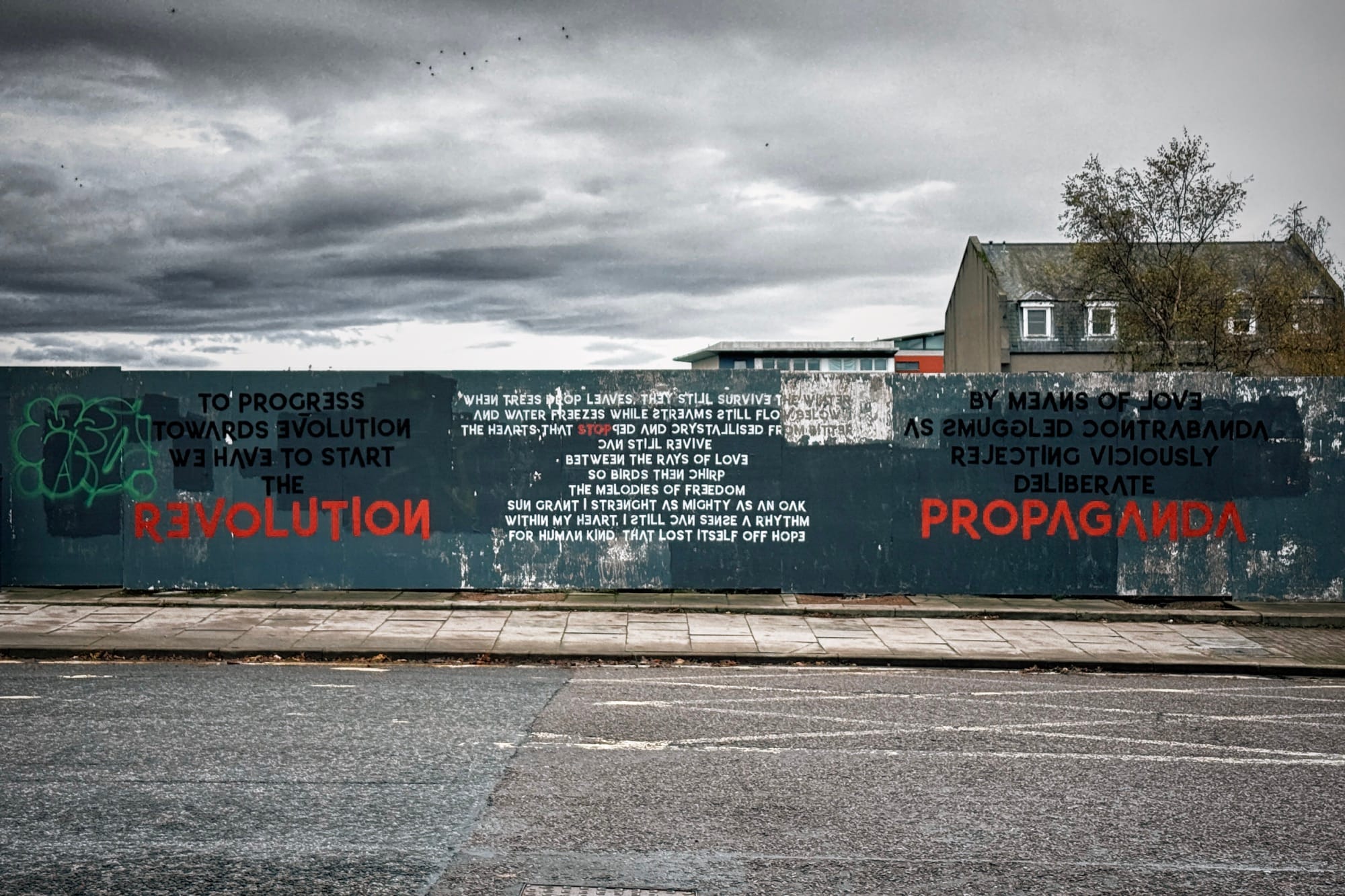 Blue wall mural with red REVOLUTION and PROPAGANDA text, poetry in centre, grey building behind.