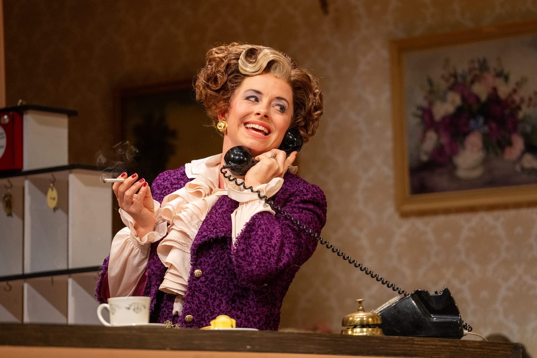 Sybil at the hotel reception, cigarette in hand, laughing into a vintage telephone