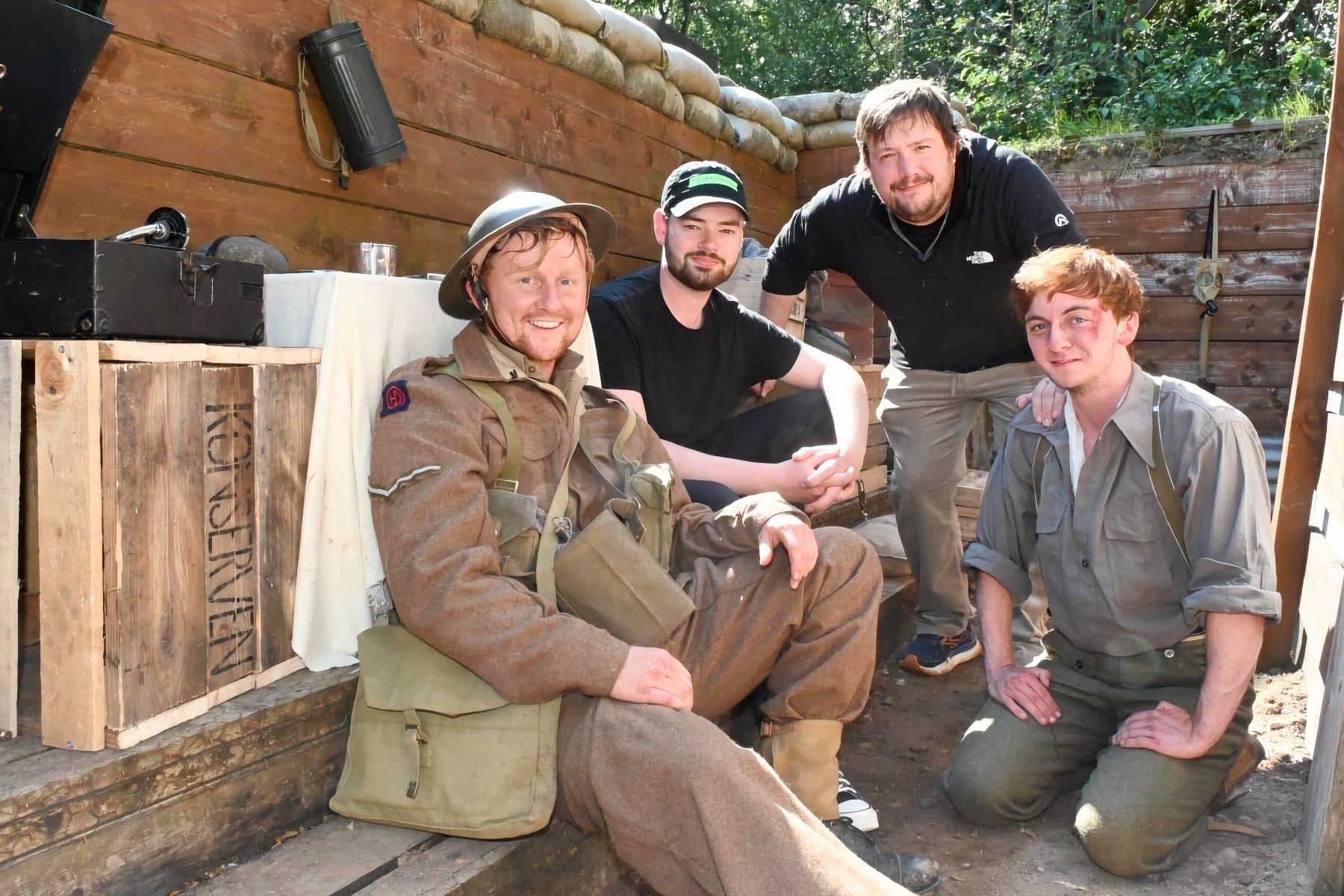 Four men posing in a recreated WWI trench set with sandbags and wooden fortifications. Two wear period military costumes while two are in modern clothing.
