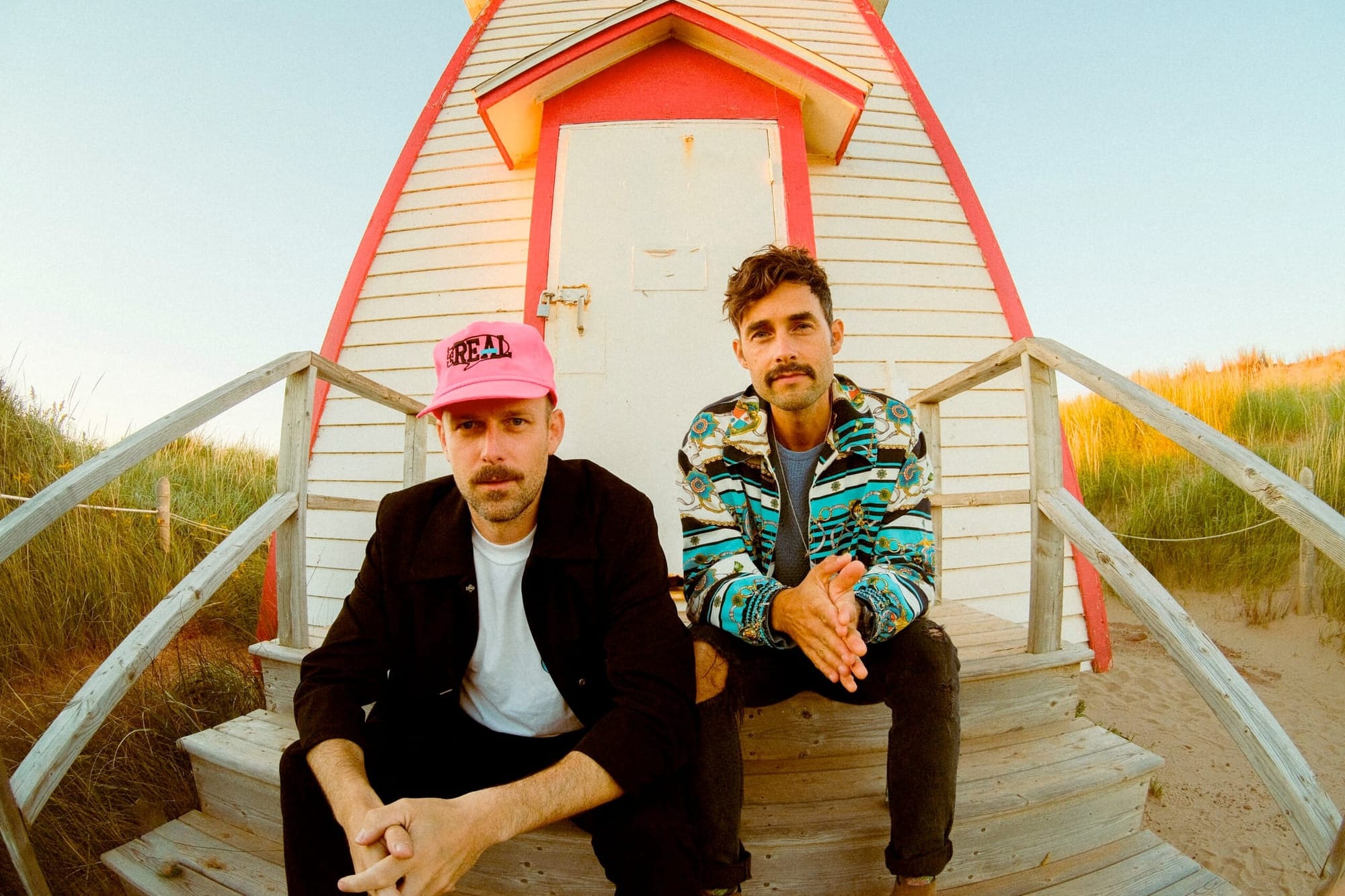 Two members of The East Pointers sitting on wooden steps in front of a beach hut with orange and cream trim, surrounded by dune grass.