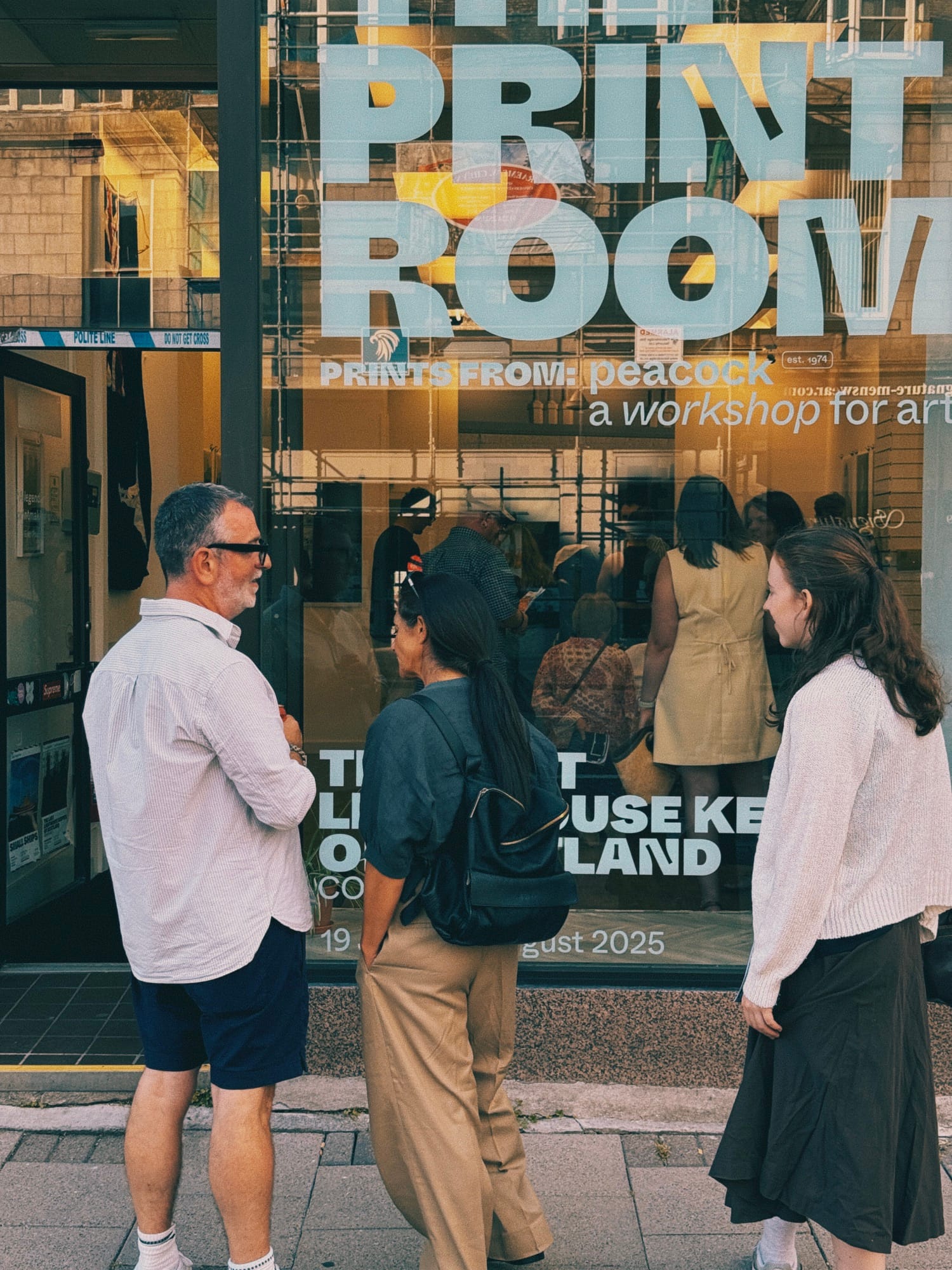 People gathered outside shopfront window displaying "PRINT ROOM" text and "prints from: peacock" signage.