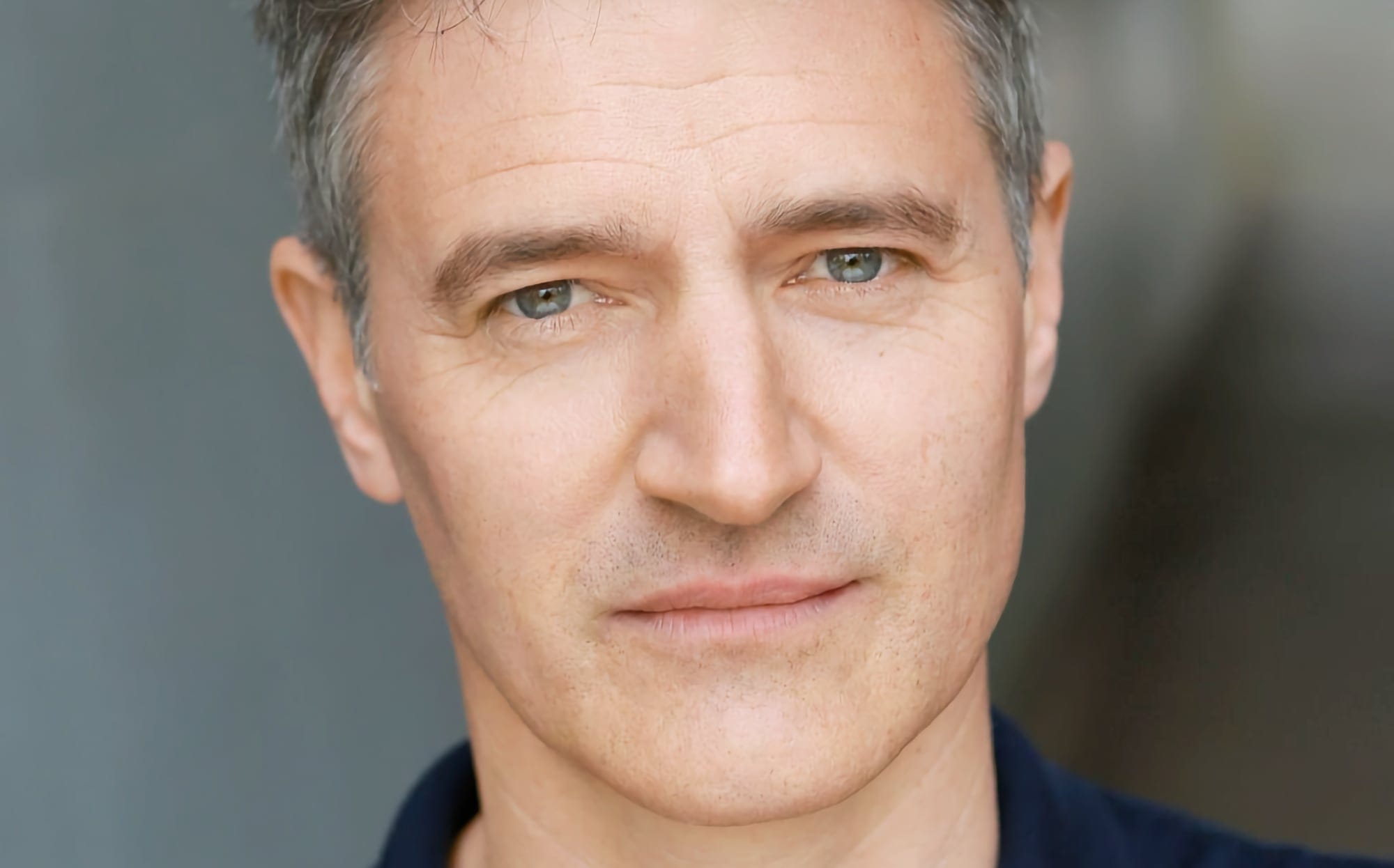 A headshot of actor Tom Chambers against a grey background, looking directly at the camera.