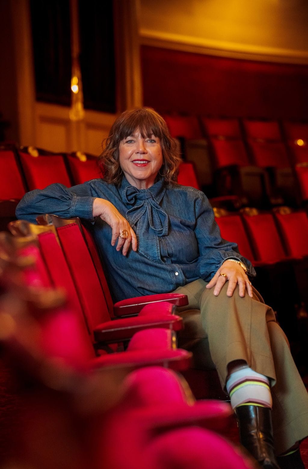 Photo of Sharon Burgess, smiling in a denim blouse, sitting in a row of red velvet theatre seats.