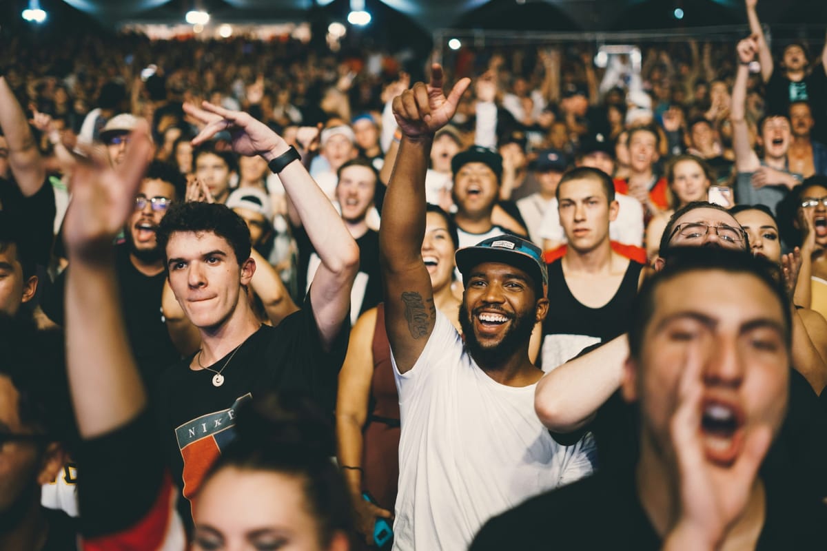 A packed crowd of people at a live event, many smiling, shouting and raising their hands in excitement. The photo captures a moment of high energy under stage lighting.
