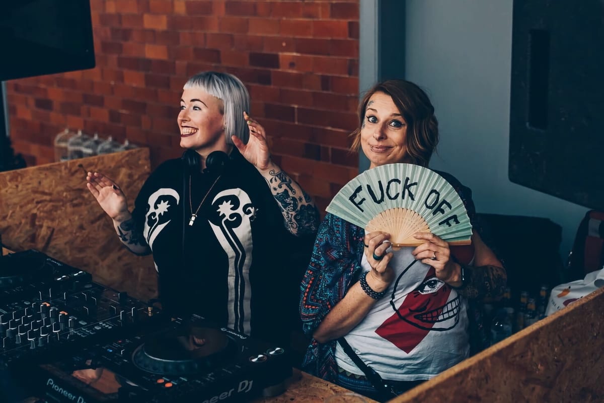 Lezzer Quest members stand smiling, one holding a “Fuck Off” fan while DJ equipment is nearby.