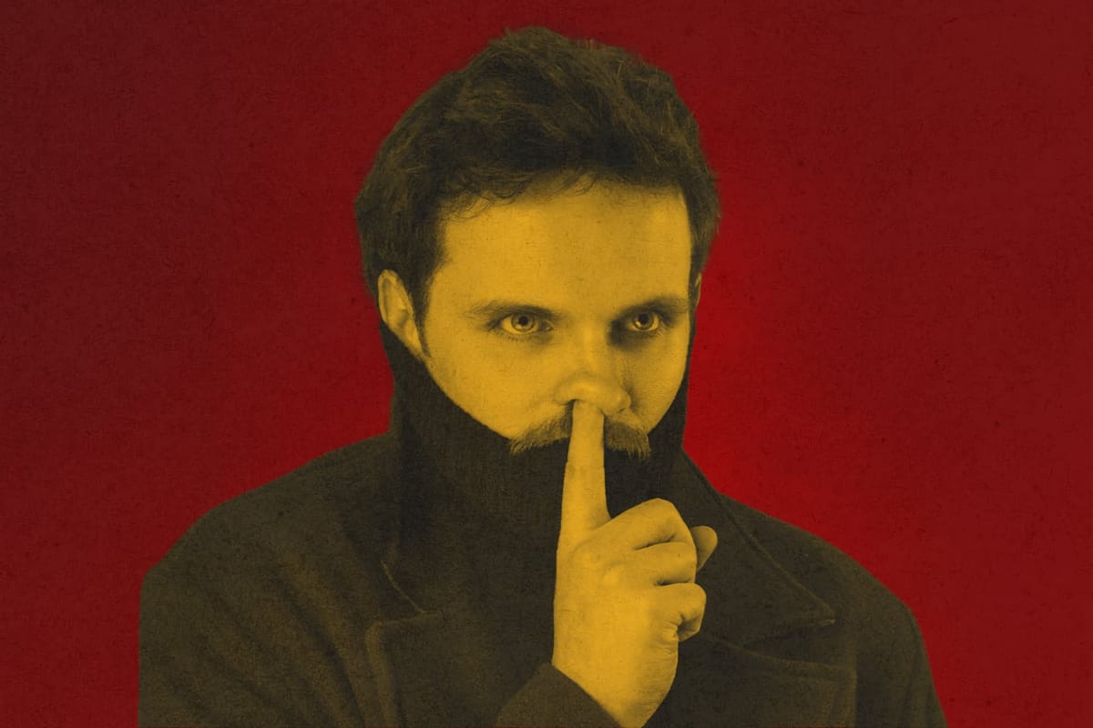 Comedian Vittorio Angelone stares ahead, finger raised to lips, set against a bold red background.