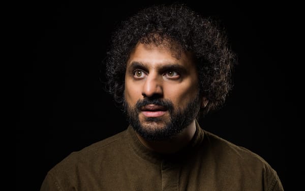The image is a professional portrait of comedian Nish Kumar, featuring him against a dark background, wearing a dark olive gr