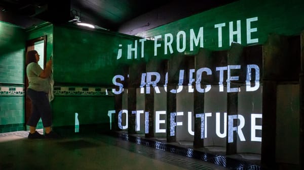 A person photographing a wall with green tiles, where the text "LIGHT FROM THE PAST PROJECTED INTO THE FUTURE" is projected.