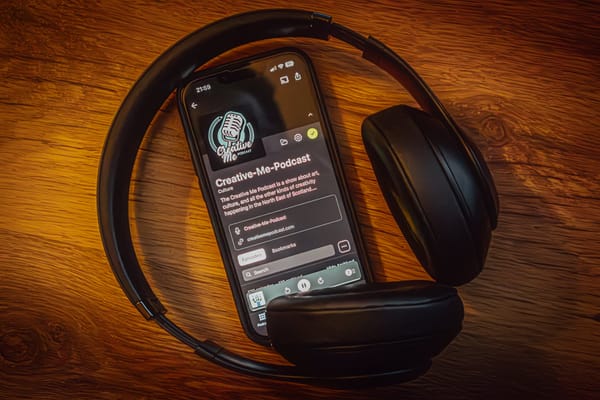 Smartphone with Creative-Me-Podcast page and logo on screen, surrounded by black headphones on a wooden surface.