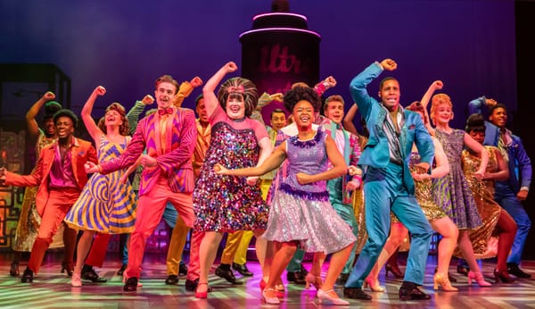 A colourful ensemble cast dances energetically in vibrant costumes on a brightly lit musical theatre stage.