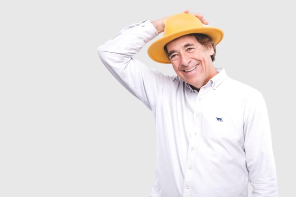 Mark Steel smiling warmly, wearing a white shirt and yellow hat, hand resting playfully on his head.