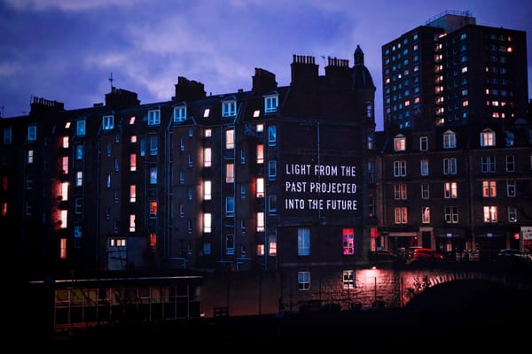 A projection on a tenement wall reads “LIGHT FROM THE PAST PROJECTED INTO THE FUTURE” as dusk settles over the city.