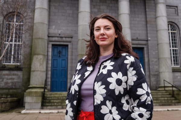 Eve Nicol stands confidently outside Aberdeen Arts Centre, wearing a black and white daisy-patterned jacket. She’s framed by the building’s grey granite columns and blue doors.