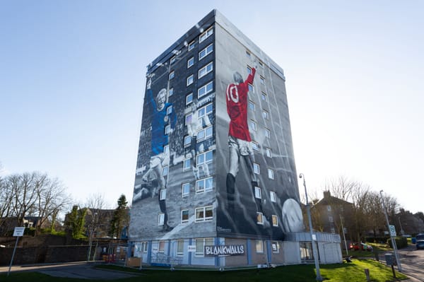Tower block mural of Denis Law in red Man Utd and blue Scotland kits, celebrating mid-match