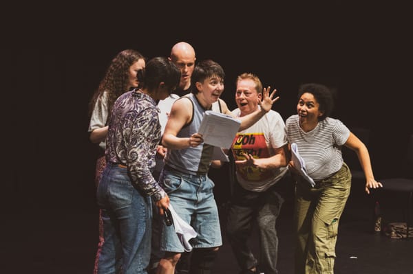 A group of performers on stage mid-rehearsal, holding scripts and reacting animatedly.