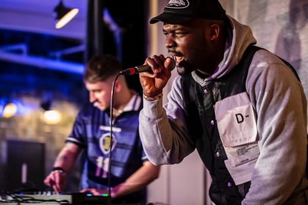 Namebliss on the mic in the foreground of a live performance, while Vagrant Real Estste DJs in the background wearing a Scotl