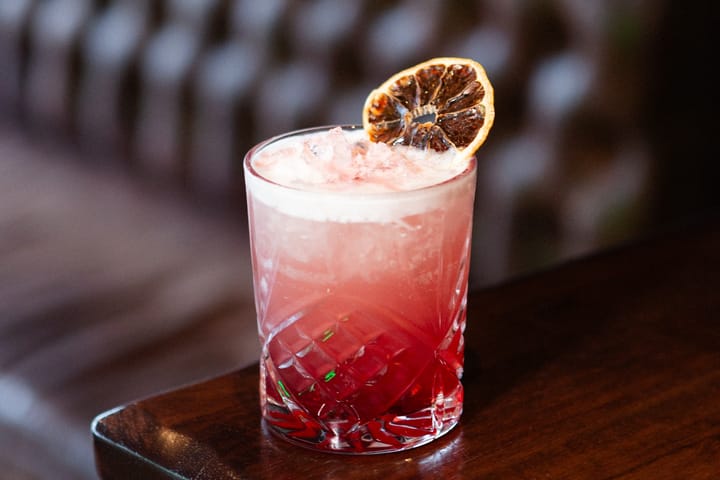 A pink cocktail in a cut-glass tumbler with ice and a dried citrus garnish on a wooden table.