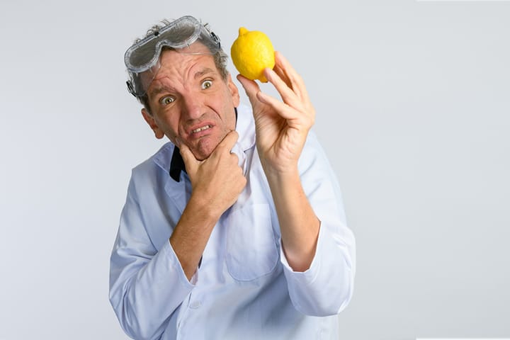 Comedian Henning Wehn in lab coat and goggles, holding a lemon with a puzzled expression.