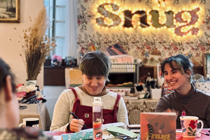 Two people chatting and drawing at a cosy craft table, with a glowing 'Snug' sign on the wall behind.