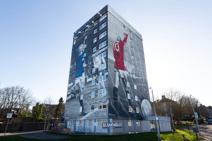 Tower block mural of Denis Law in red Man Utd and blue Scotland kits, celebrating mid-match