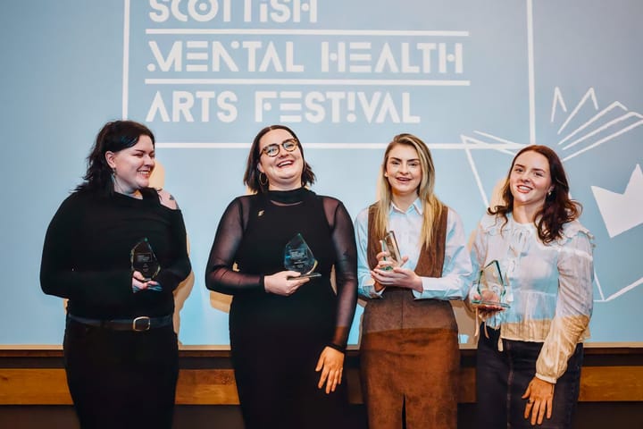 Four women holding glass awards standing together smiling in front of a Scottish Mental Health Arts Festival backdrop.