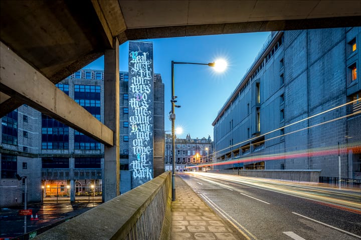 A tall vertical mural on a grey building displaying ornate white calligraphic text, photographed at dusk from a road bridge with light trails from passing traffic visible in the foreground.