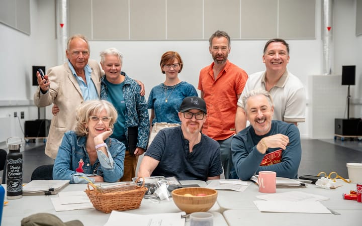 Cast and creative team at a table in a rehearsal room with scripts and mugs