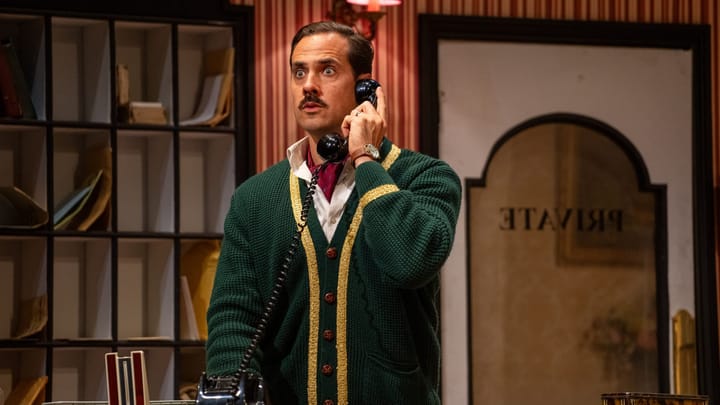 Basil on the phone at reception, eyes wide with alarm, wearing a green cardigan
