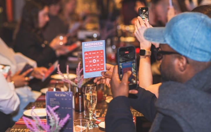 Diners at a restaurant table photographing the Aberdeen Restaurant Weeks bingo card