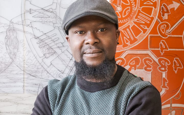 Artist Ade Adesina wearing a flat cap, standing in front of his detailed line drawings.