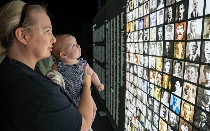 Woman holding a baby looks at a grid of Titanic passenger photographs on a dark wall.