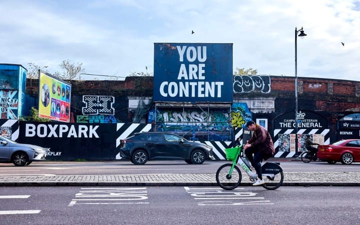 A cyclist passes a billboard reading "You Are Content" on a busy urban street.
