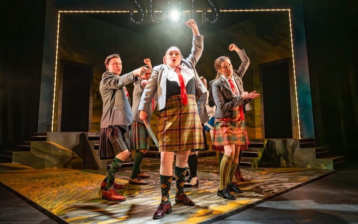 Ensemble cast in kilts and school uniforms perform on stage with fists raised