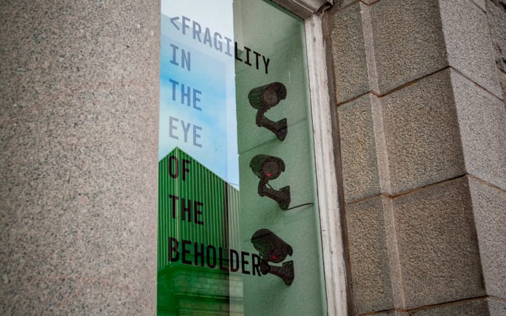 Three CCTV cameras in a shopfront window beside a poster reading Fragility in the Eye of the Beholder