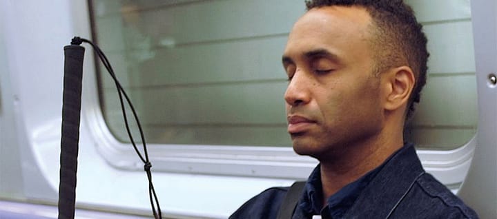 Vision Portraits director Rodney Evans sits on an underground train with the window behind him. His eyes are closed and the b